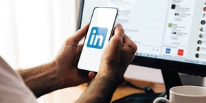 Hands holding smart phone with LinkedIn logo on it and computer blurred in the background with the LinkedIn application open.